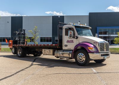Elite Transportation Systems flatbed truck with a piggyback forklift on the rear, parked outside a warehouse