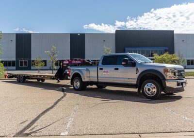 Elite Transportation Systems pickup with an empty gooseneck flatbed trailer outside a commercial building