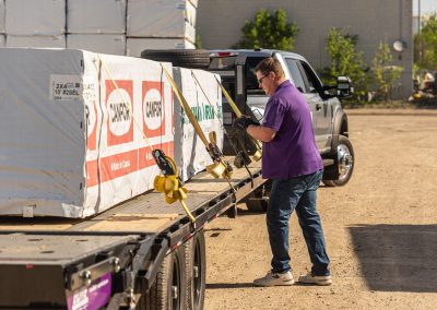 Elite Transportation Systems contract driver tightening a ratchet strap over wrapped lumber on a gooseneck flatbed